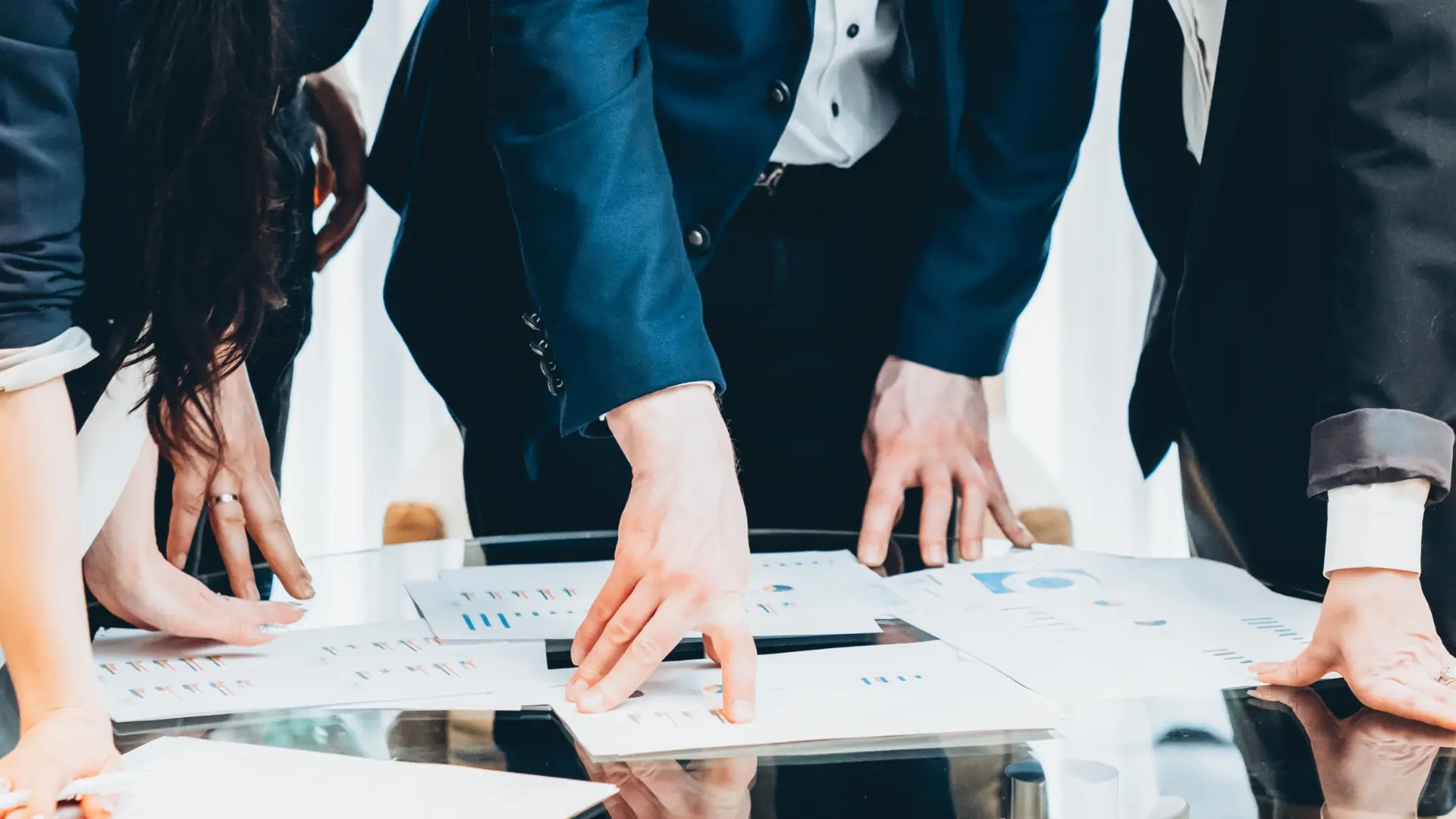 Business professionals in formal attire reviewing and pointing at charts and graphs on documents laid out on a glass table, indicating a collaborative financial analysis or strategic planning session