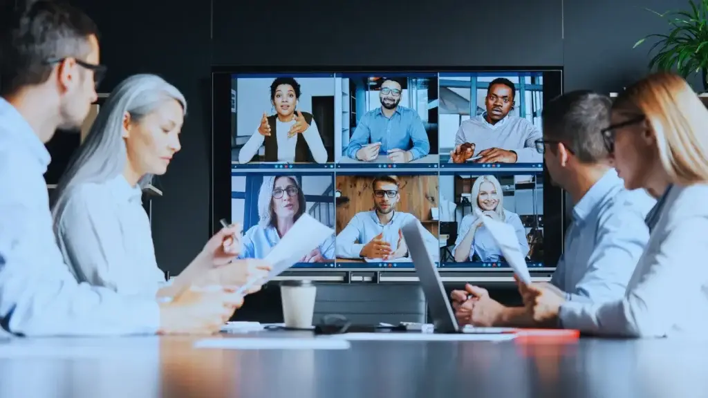 Professional team in a hybrid meeting with colleagues on a video call displayed on monitor, engaging in corporate strategy session, blending in-person and virtual collaboration.
