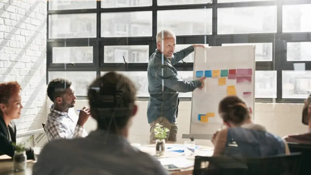 Casual meeting with a diverse group focused on a man explaining sticky notes on a whiteboard.