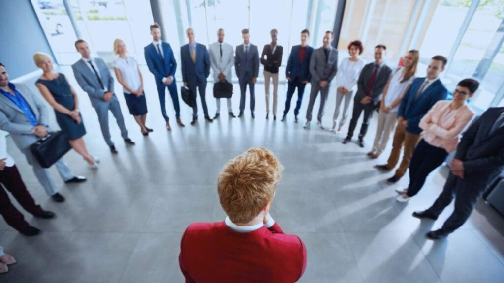 Rear view of a leader in a red jacket addressing a diverse group of professionals standing in a semicircle in a bright office setting.