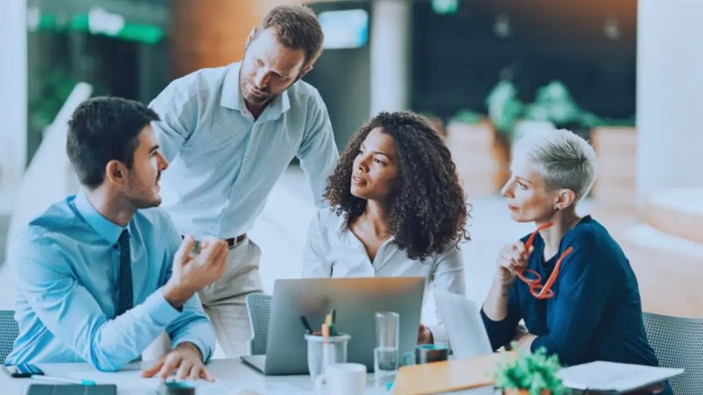 Diverse business team engaged in discussion at a modern office, with a Caucasian man standing and explaining a point to a seated group including an African American woman and two Caucasian colleagues.