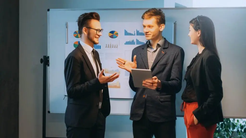 Three business professionals discussing statistical data on a digital tablet in front of a whiteboard with charts and graphs, illustrating the salience model in an office setting.