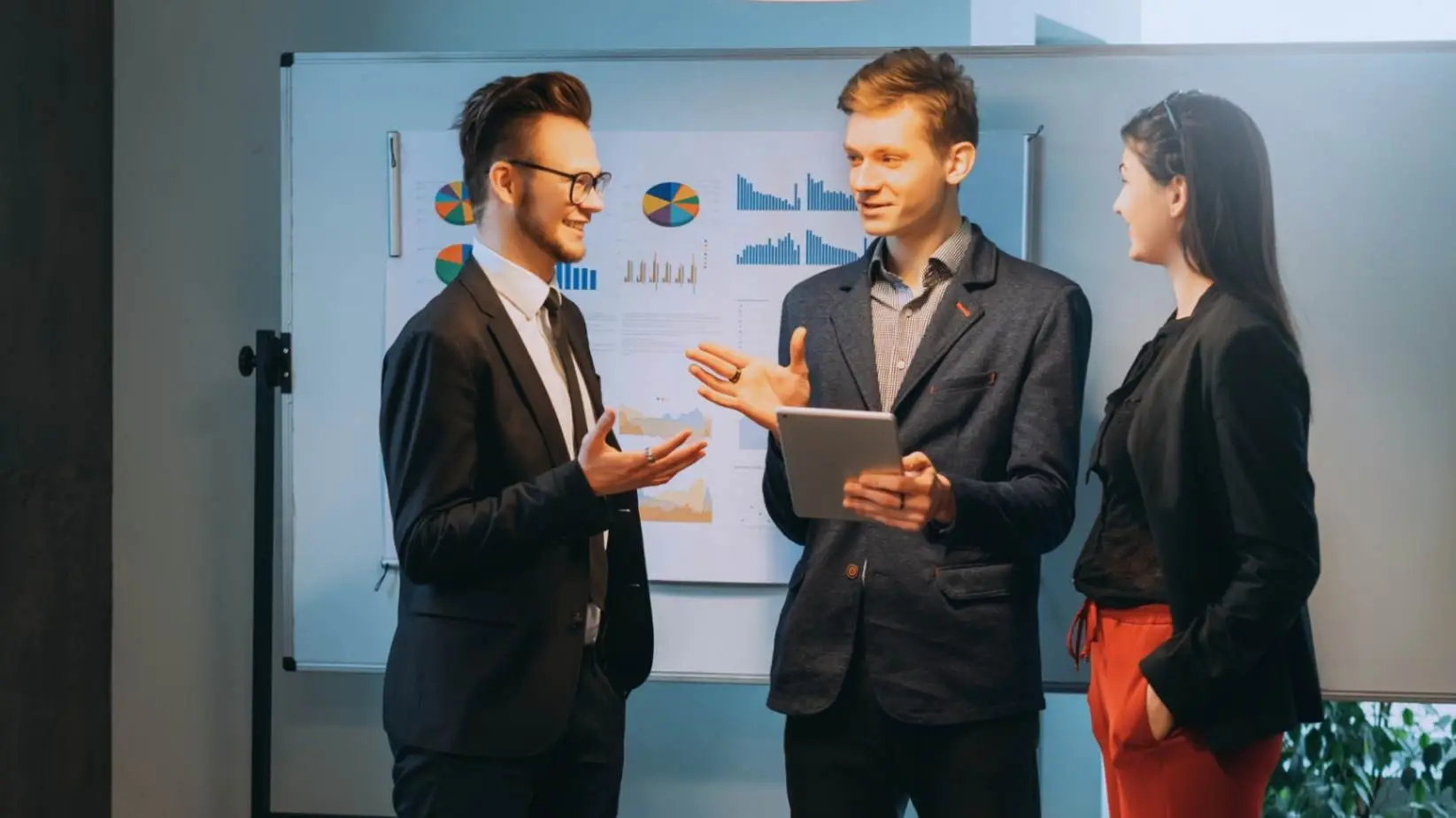 Three business professionals discussing statistical data on a digital tablet in front of a whiteboard with charts and graphs, illustrating the salience model in an office setting.