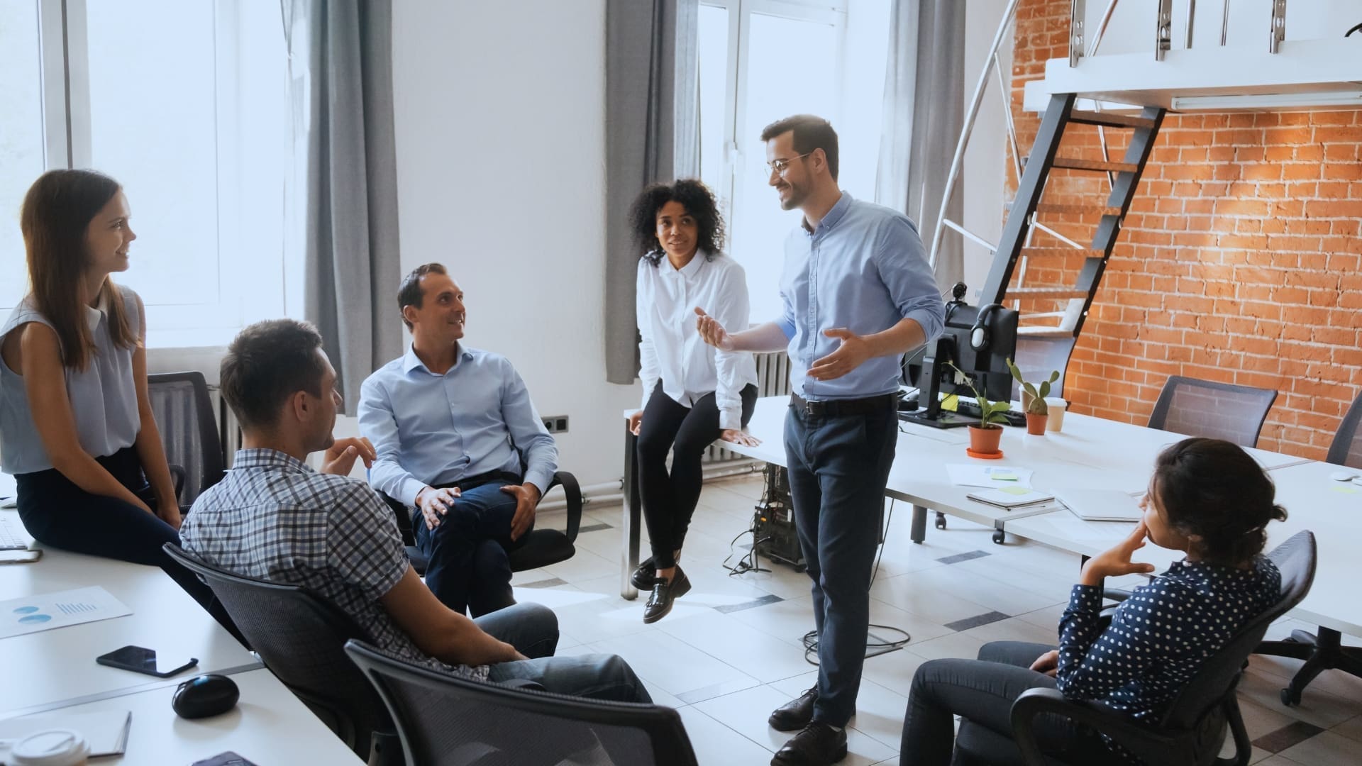 A group of six people in a casual office setting have a meeting. Four people, three men and one woman, sit while one man and one woman stand. There is a bright window, a brick wall, and various office furniture, including desks and chairs, in the background. Leadership principles guide their discussion.