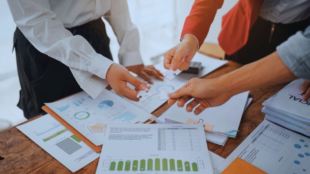 Several individuals collaborate at a wooden table, handling puzzle pieces while surrounded by graphs, charts, documents, and project charters. The image depicts a collaborative business or team planning environment with hands actively engaged in discussion and problem-solving.