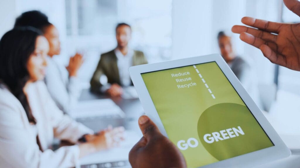 A person holds a tablet displaying the message "Reduce, Reuse, Recycle" and "Go Green" in front of a group of people seated at a conference table, engaged in a discussion about sustainability in project management. The room is well-lit with natural light.