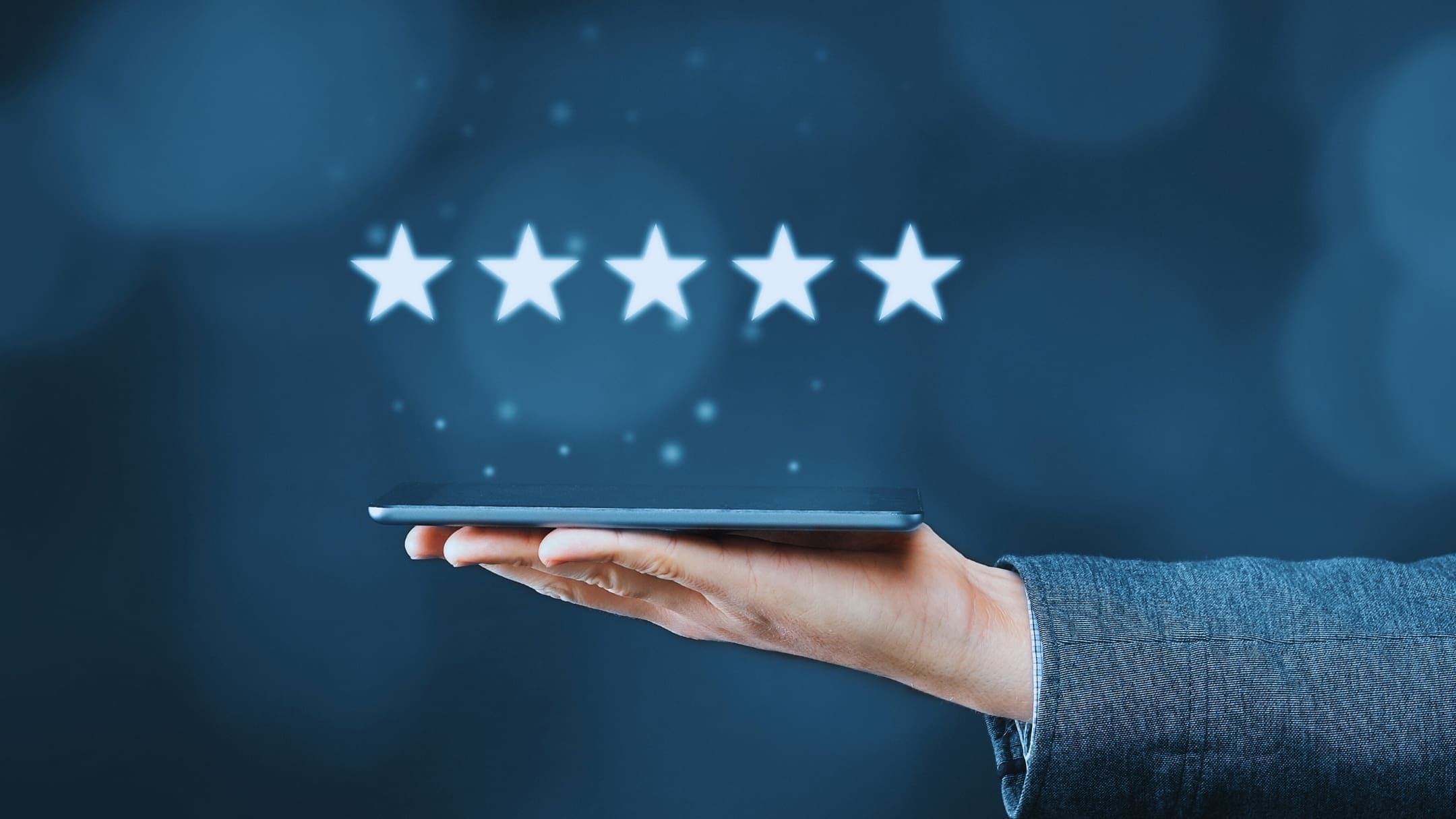 A hand in a suit holds a horizontal tablet with five white stars floating above it against a dark blue, bokeh background, subtly questioning, "Is Notion a good app?" while representing a five-star rating.
