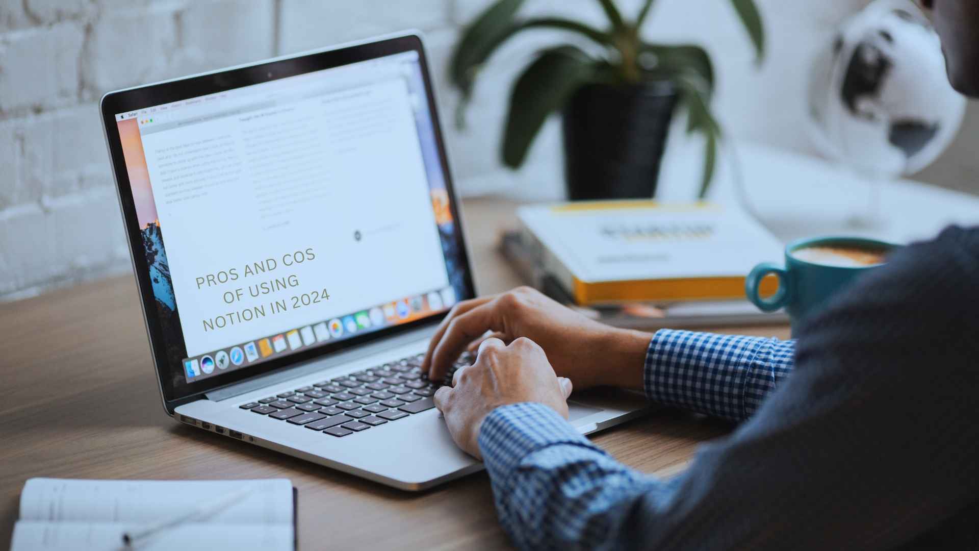 A person wearing a checkered shirt is typing on a laptop at a wooden desk. The laptop screen displays a document titled "Pros and Cons of Using Notion in 2024," evidently working on a notion software review. There is a plant, a book, and a blue mug on the desk.