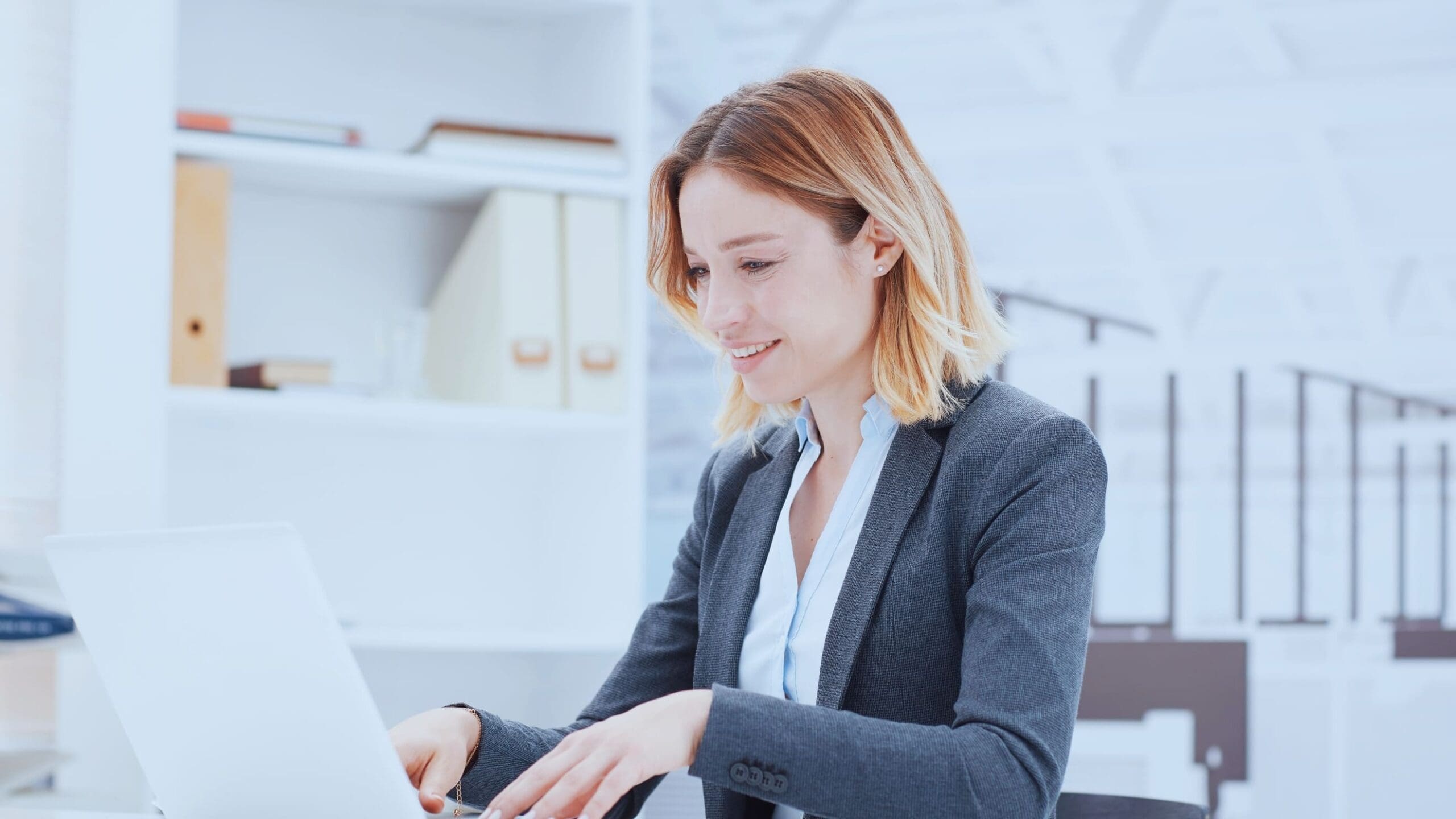 Professional woman smiling while working on a laptop in a modern office environment.