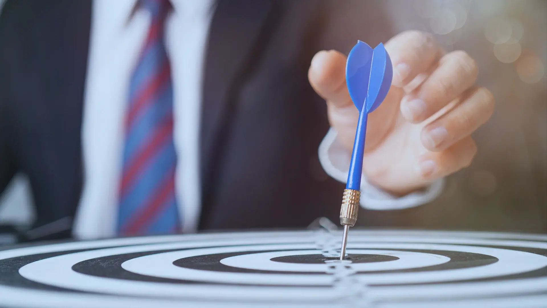 A person in a suit and tie holds a blue dart above a dartboard, aiming to hit the bullseye—an image that represents focus, goal setting, and achieving objectives.