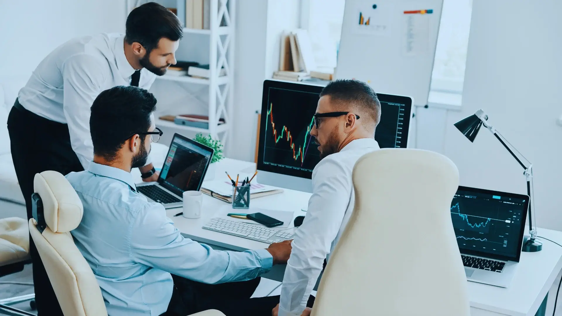 Three men in business attire work together at a desk with computers displaying financial charts and graphs, suggesting a stock market meeting focused on quantifying risk and financial analysis in a modern office.