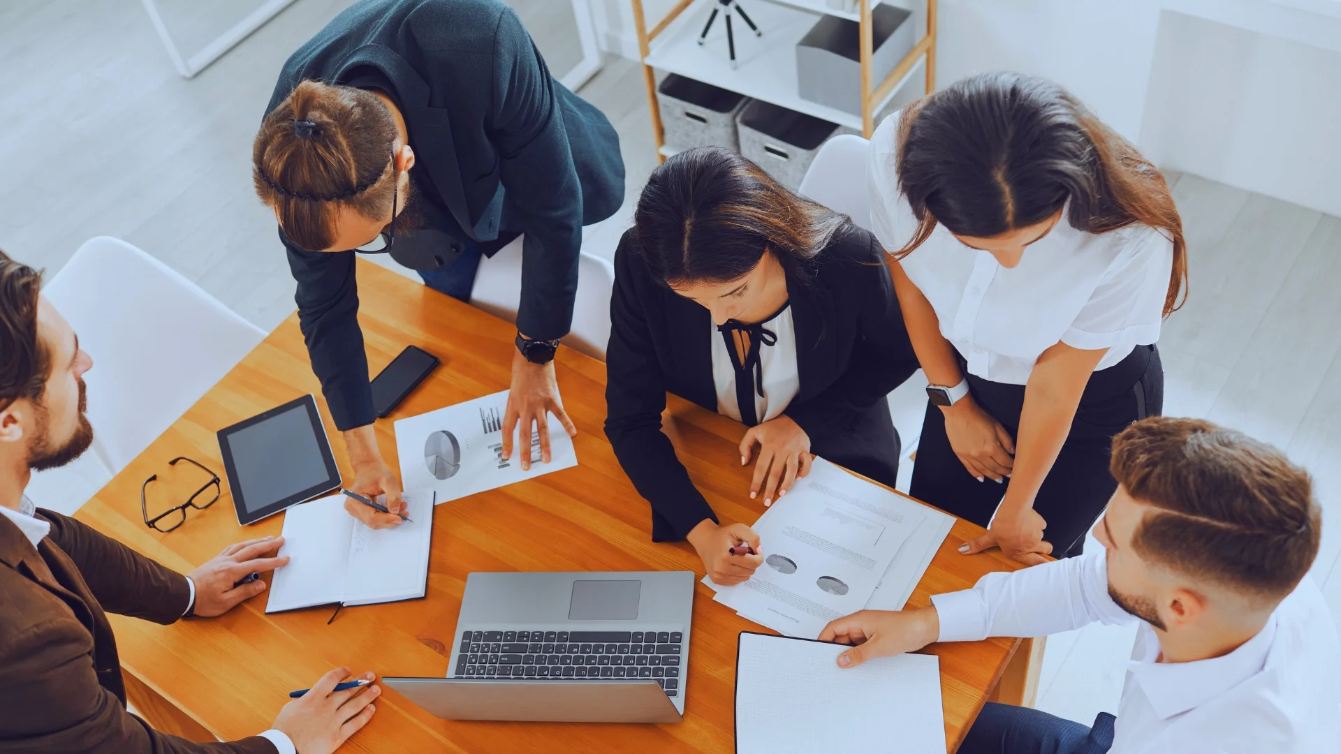Five business professionals in formal attire are gathered around a wooden table, collaborating over documents, charts, and a laptop as they focus on quantifying risk in a bright office setting.