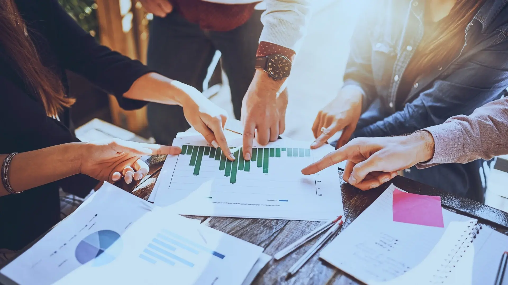 Several people’s hands point at a bar graph on a paper among scattered documents and pens on a wooden table, suggesting a collaborative meeting focused on quantifying risk or analyzing business data.