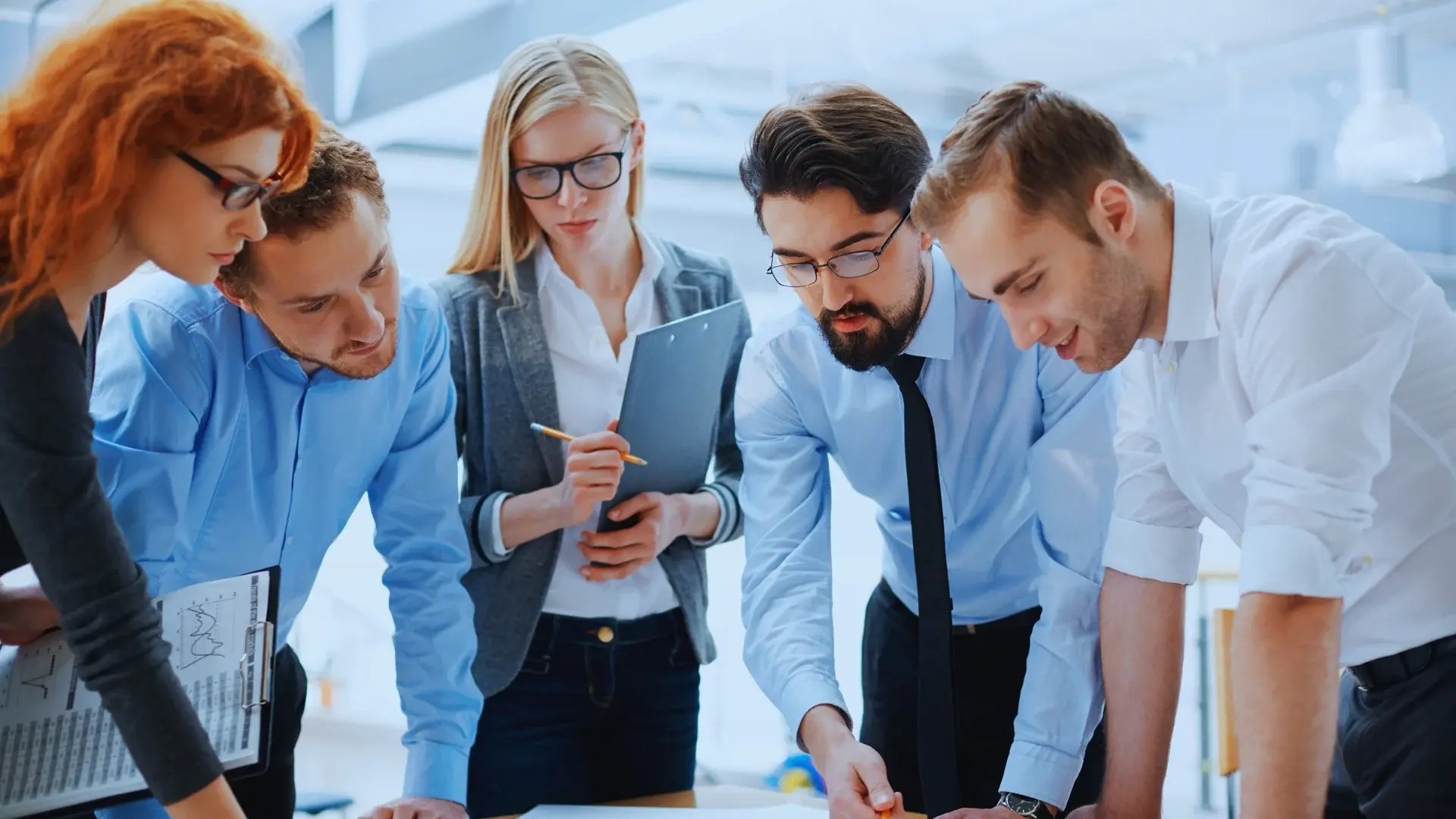 Four men and one woman in business attire gather around a table, reviewing documents and discussing work. The woman holds a clipboard as the group analyzes papers, suggesting a collaborative office meeting focused on quantifying risk.