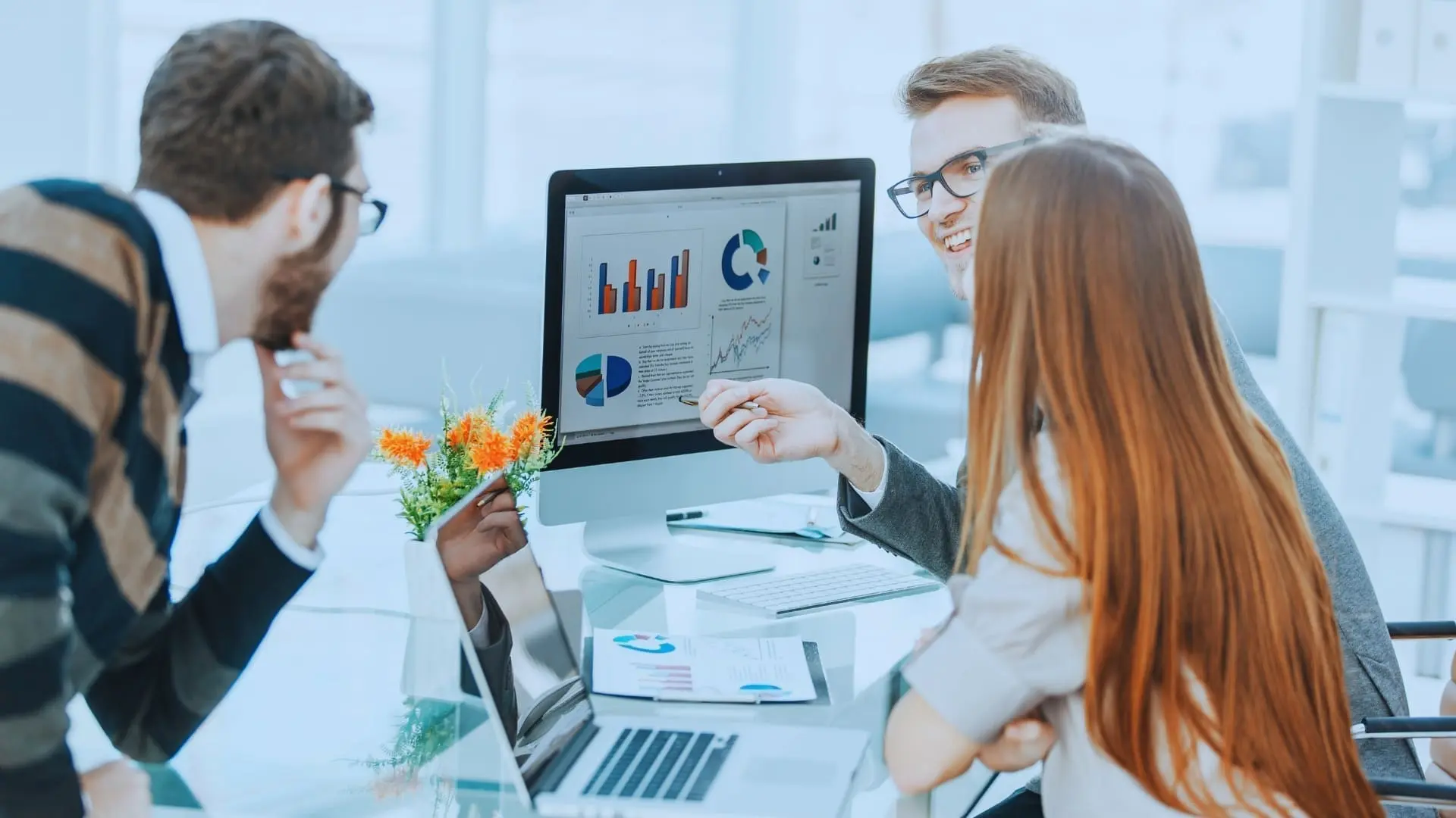 Three colleagues sit around a desk, analyzing charts and graphs comparing BVDash vs SmartSuite on a computer screen. One person points at the display as they engage in a focused business meeting discussion.