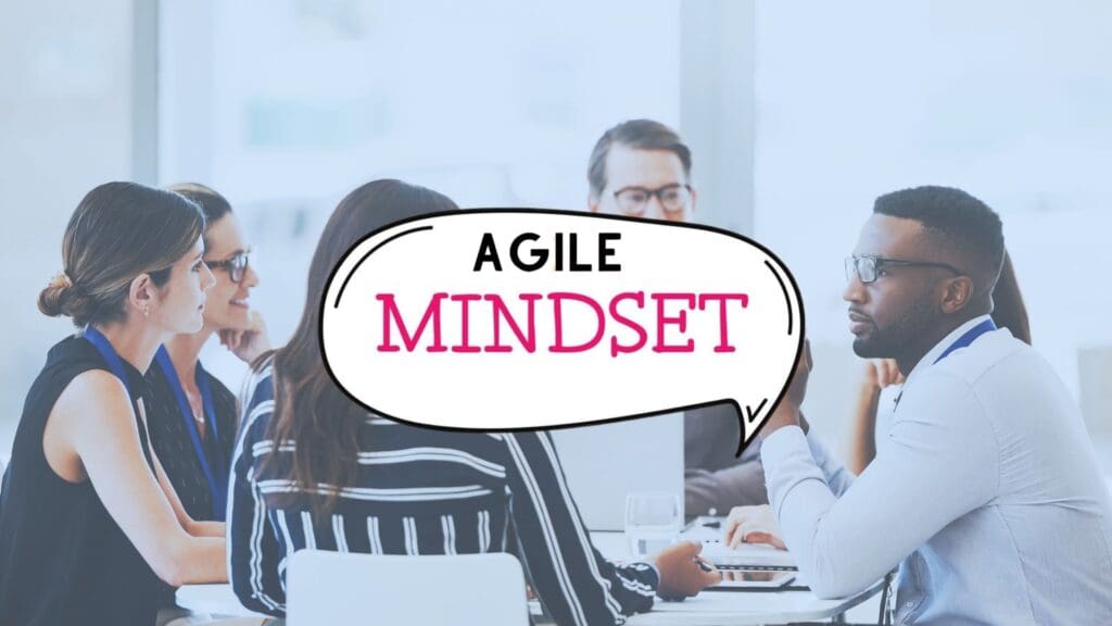 A group of people engaging in a discussion around a table in a modern office setting. A speech bubble in the center reads "AGILE MINDSET" with the word "MINDSET" highlighted in pink. They all appear focused and are likely part of a collaborative Agile meeting.