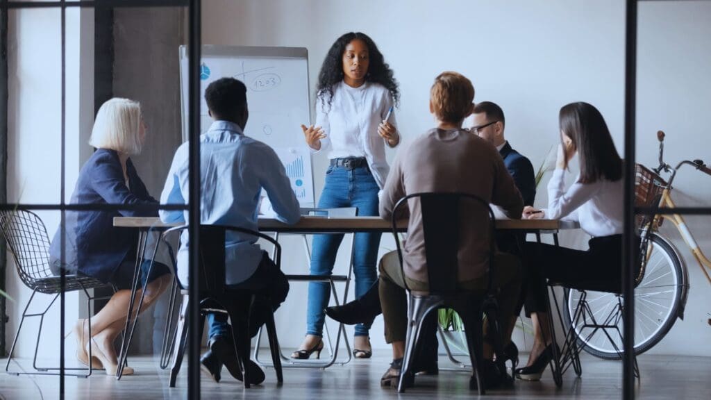 A woman, acting as the team facilitator, stands and presents to a group of five seated individuals in a modern office setting. She gestures towards a whiteboard with various diagrams. The group appears engaged, with a bicycle and large windows in the background adding to the contemporary vibe.