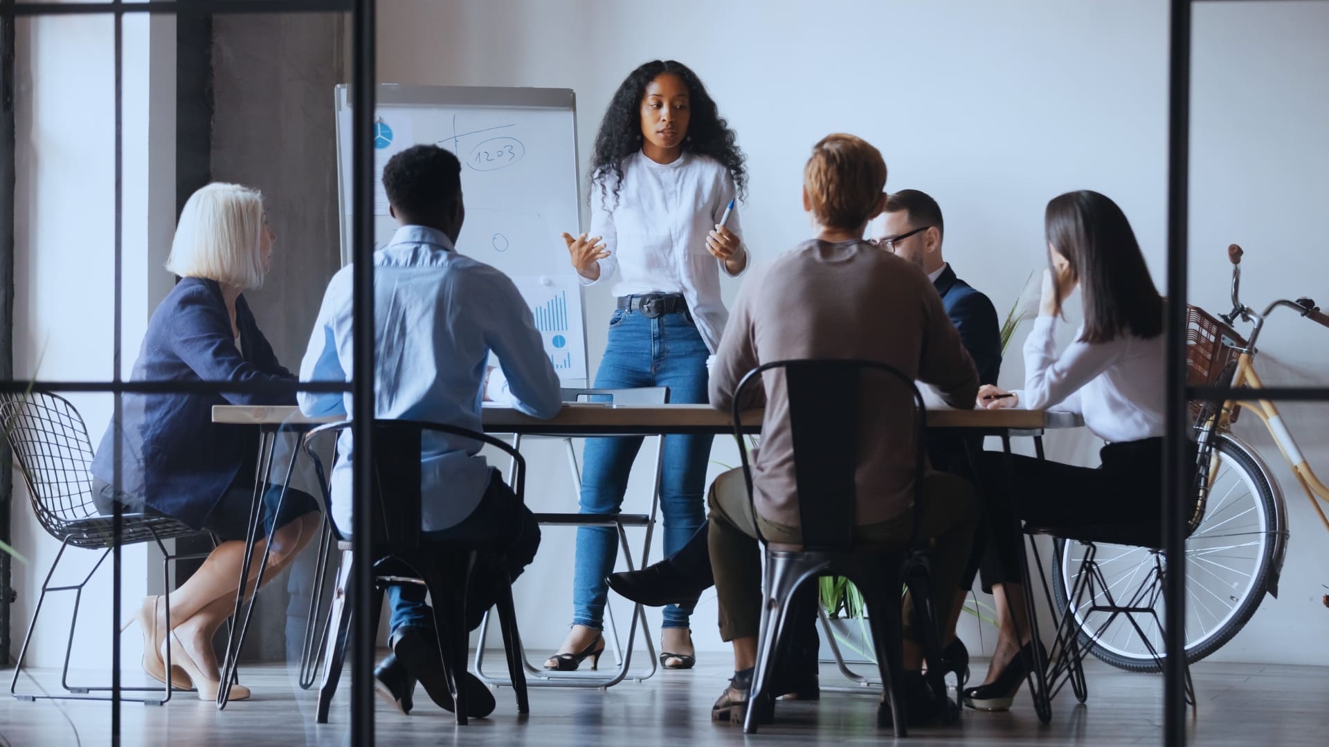 A woman, acting as the team facilitator, stands and presents to a group of five seated individuals in a modern office setting. She gestures towards a whiteboard with various diagrams. The group appears engaged, with a bicycle and large windows in the background adding to the contemporary vibe.