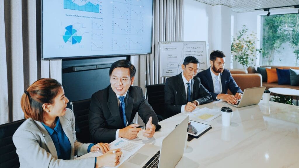 Four businesspeople in a conference room are having a TeamGantt review meeting. Three are seated with laptops in front of them, while the fourth person is gesturing during the discussion. There is a screen displaying charts and graphs and a whiteboard with diagrams in the background.