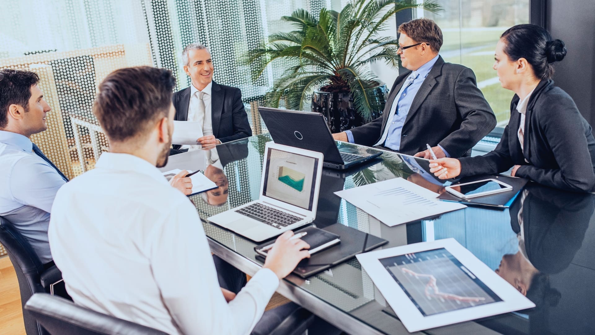 Five people in formal business attire are sitting around a glass conference table engaged in a meeting. Two laptops and documents with graphs and charts are on the table, likely discussing business strategies or financial data. They're exploring ways to enhance productivity, possibly considering free monday.com tools.