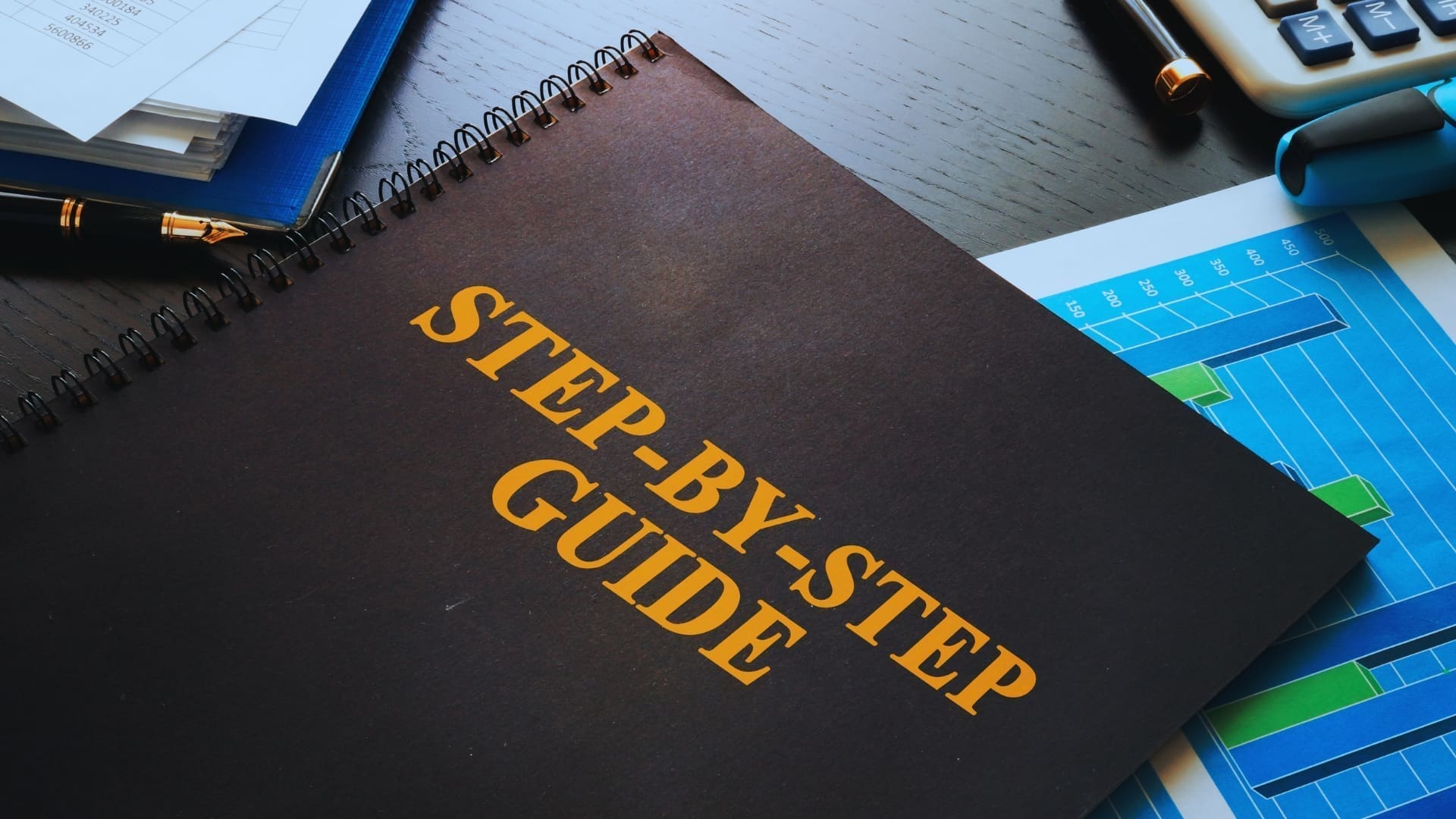 A black notebook with the title "STEP-BY-STEP GUIDE" in bold yellow letters sits on a wooden desk. Surrounding it are a blue pen, a graph printout in shades of blue and green, a calculator, and other papers, ready to help you boost productivity after your monday.com sign up.