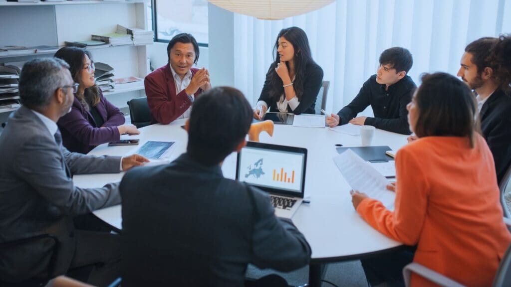 In a modern office, eight people are having a meeting around a circular table. Laptops with charts, papers, and Monday.com dashboards are spread out on the table. One person is leading the discussion while the others listen and take notes. Shelves in the background hold files and office supplies.