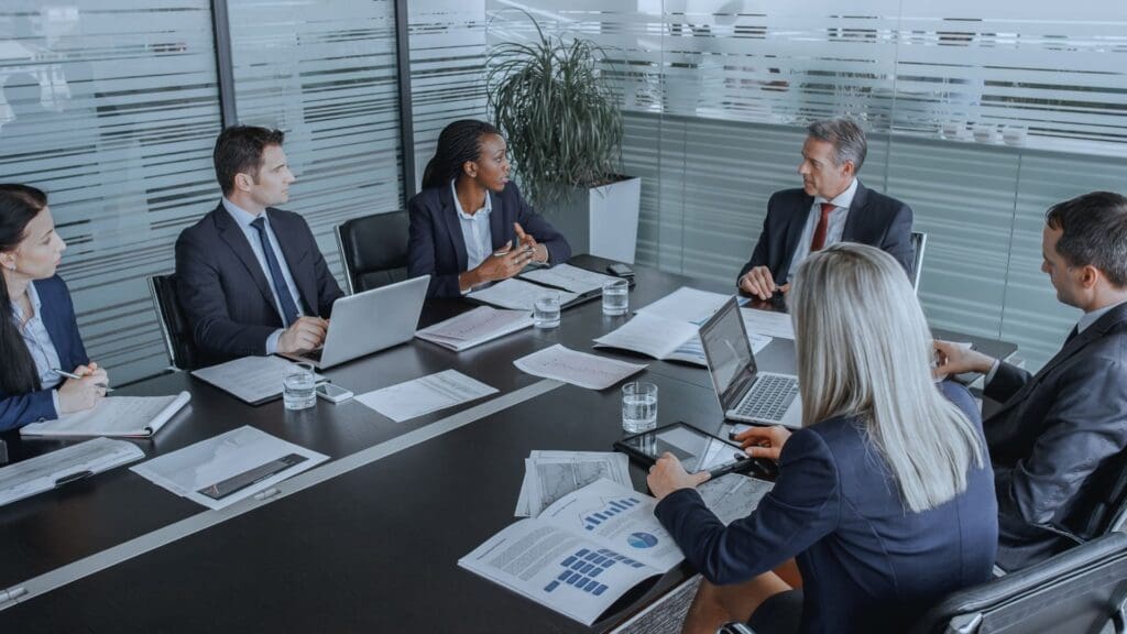 Six business professionals in formal attire are gathered around a conference table in a glass-walled meeting room. They are engaged in a discussion about TeamGantt vs. Wrike, with laptops, documents, and charts spread out on the table. A potted plant is visible in the background.