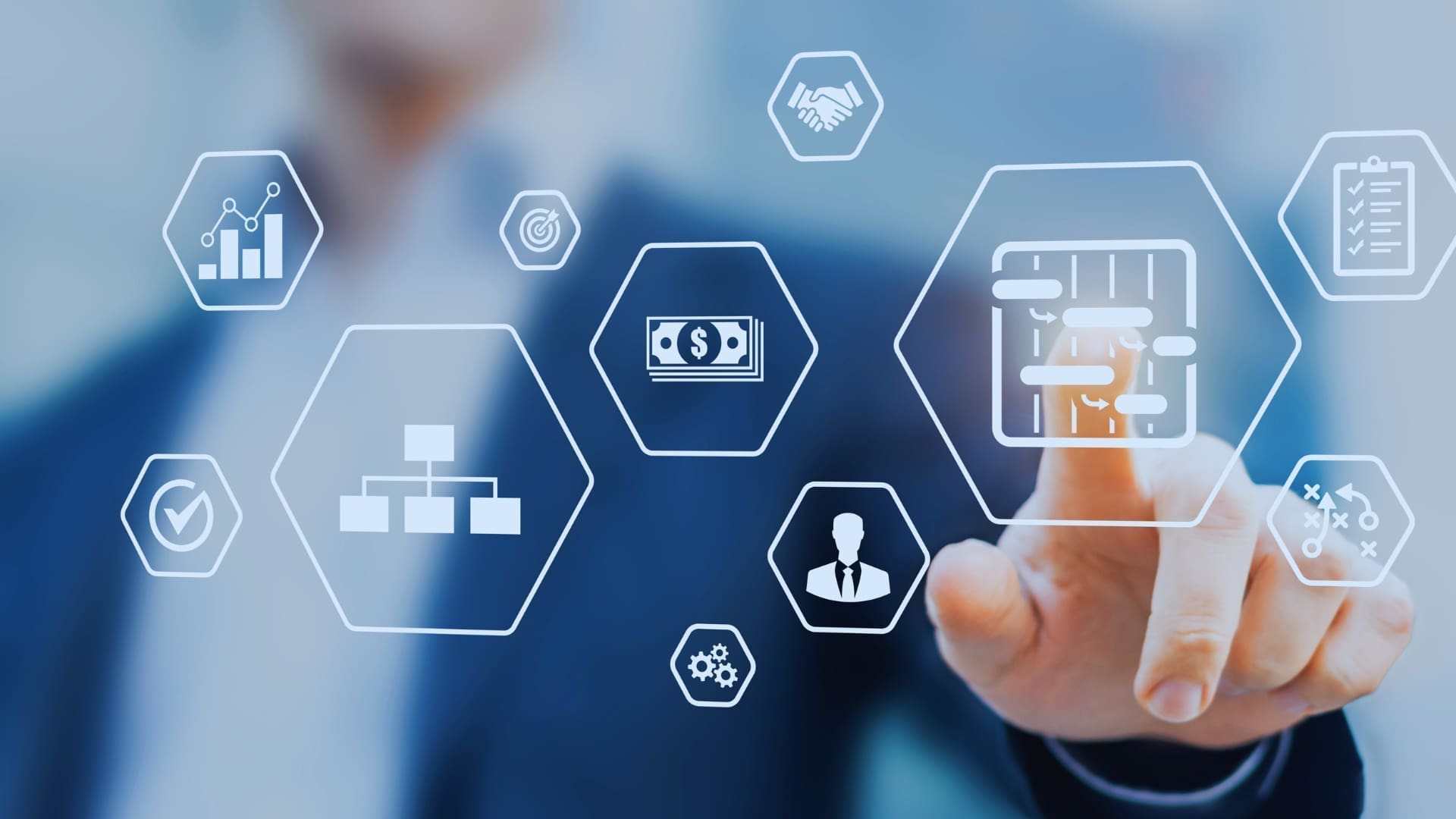 A person in a suit is touching a virtual screen with hexagonal icons depicting various business-related topics such as charts, money, organizational structure, handshake, and a person. The blurred background emphasizes the icons and hand interaction, reminiscent of comparing tools like TeamGantt vs Monday.com.