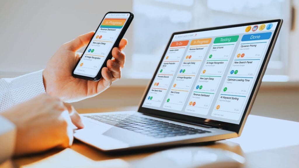 A person is using a laptop and holding a smartphone, both displaying a digital task management board in kanban view. The board on both devices shows various columns such as "To Do," "In Progress," "Testing," and "Done," with colorful task cards under each column.