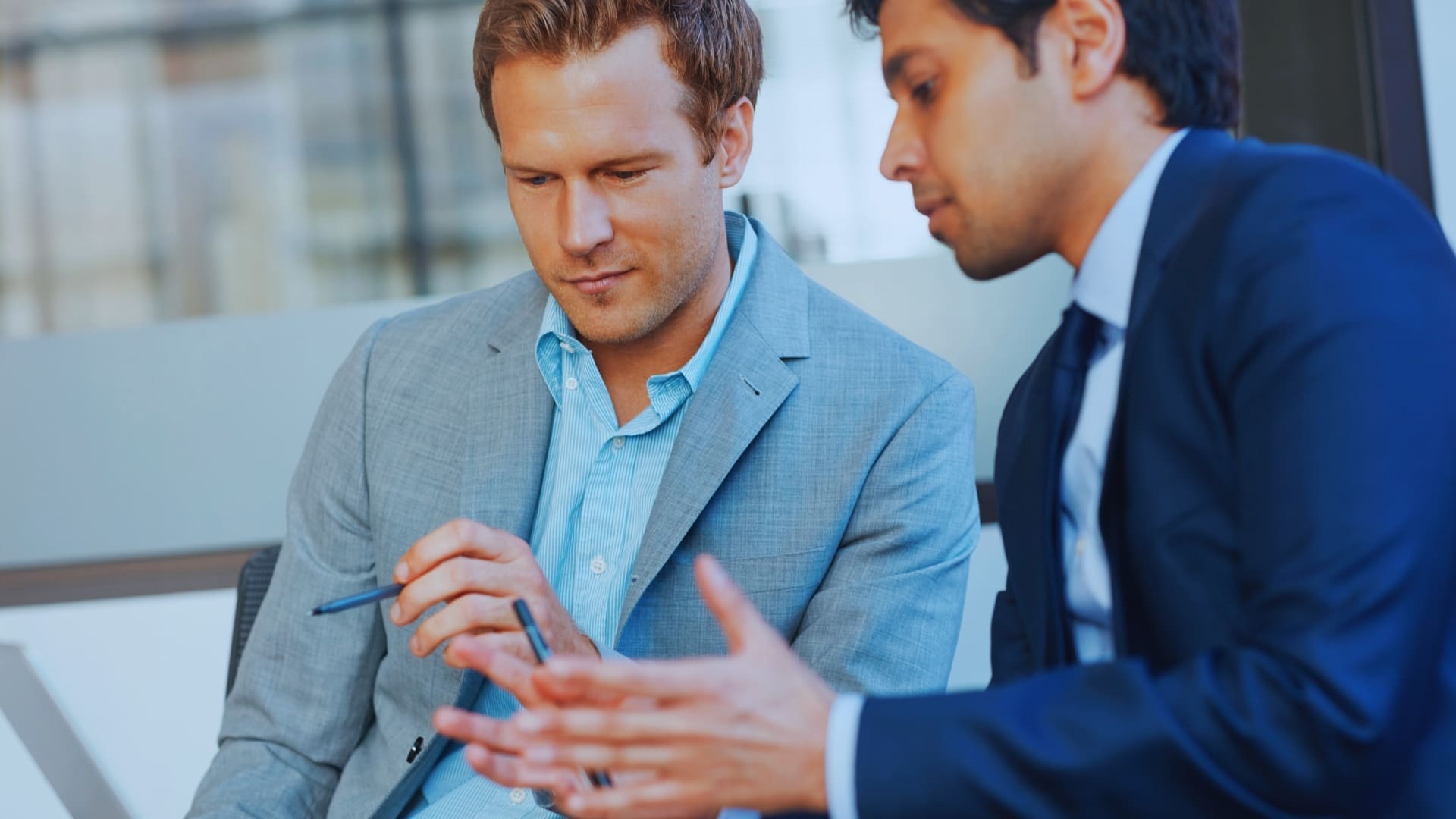 Two men in business attire engage in a discussion. One man in a light grey suit gestures with a pen, while the other man in a dark suit, referencing Monday.com's price, explains something using expressive hand movements. They appear to be in a professional setting.