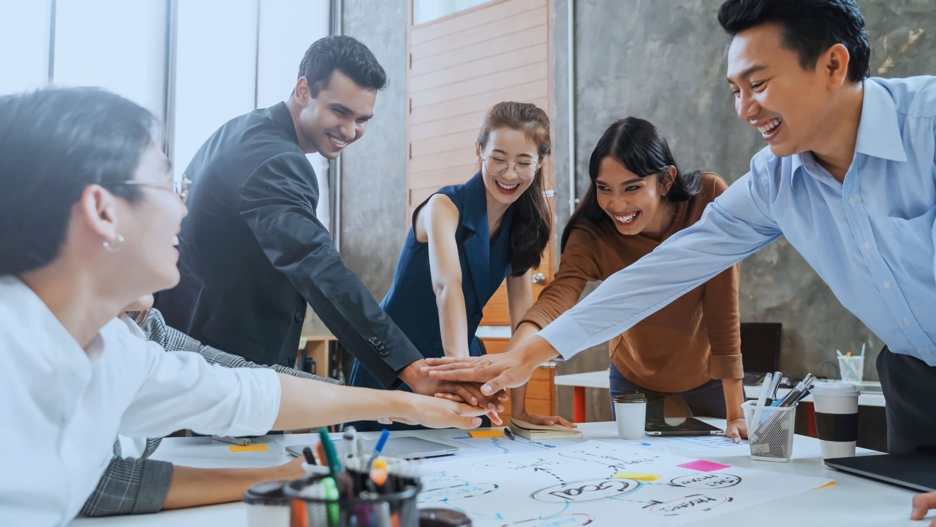 A group of five people stand around a table, placing their hands together in a gesture of unity. They are smiling and appear to be in an enthusiastic and collaborative mood, exemplifying the spirit of motivating teams. The table is covered with papers, diagrams, and office supplies.