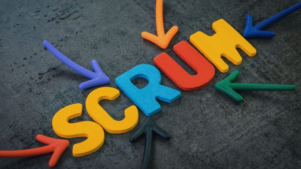 The word "SCRUM" is spelled out in colorful, 3D foam letters on a dark, textured background. Arrows in various colors point towards the word from different directions, illustrating its centrality and importance. This visual invites the question: What is Scrum in Agile?
