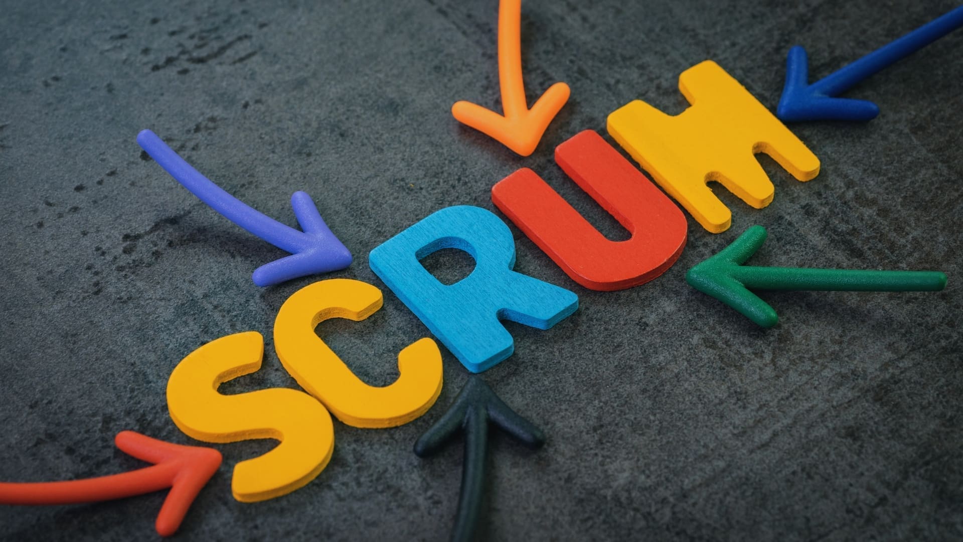 The word "SCRUM" is spelled out in colorful, 3D foam letters on a dark, textured background. Arrows in various colors point towards the word from different directions, illustrating its centrality and importance. This visual invites the question: What is Scrum in Agile?