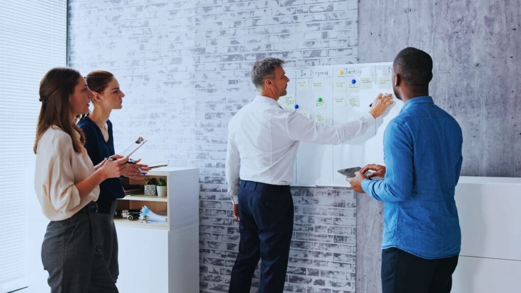 Four colleagues collaborate in an office, immersed in agile planning in agile. Two women and two men stand near a whiteboard. One man writes notes on sticky notes categorized by 'In Progress' and 'To-Do'. The others hold tablets, engaging in discussion. The backdrop features a brick wall.