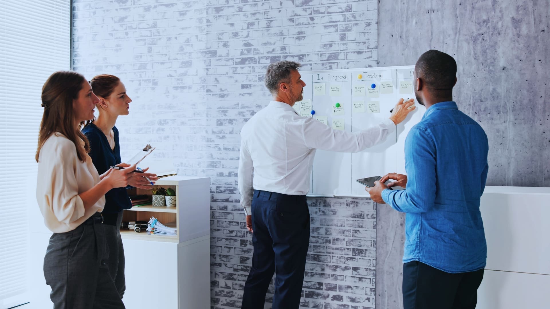 Four colleagues collaborate in an office, immersed in agile planning in agile. Two women and two men stand near a whiteboard. One man writes notes on sticky notes categorized by 'In Progress' and 'To-Do'. The others hold tablets, engaging in discussion. The backdrop features a brick wall.