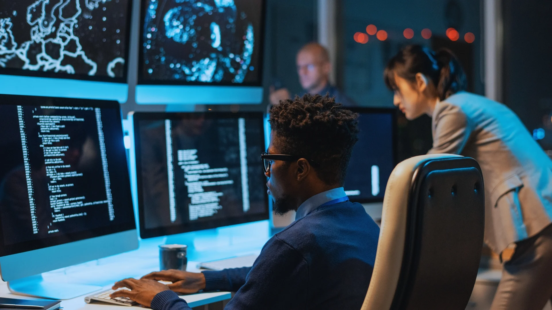 A man sits at a desk working on code related to quantifying risk, with data and graphics displayed across multiple monitors, while two colleagues work in the background of a modern office with blue lighting.