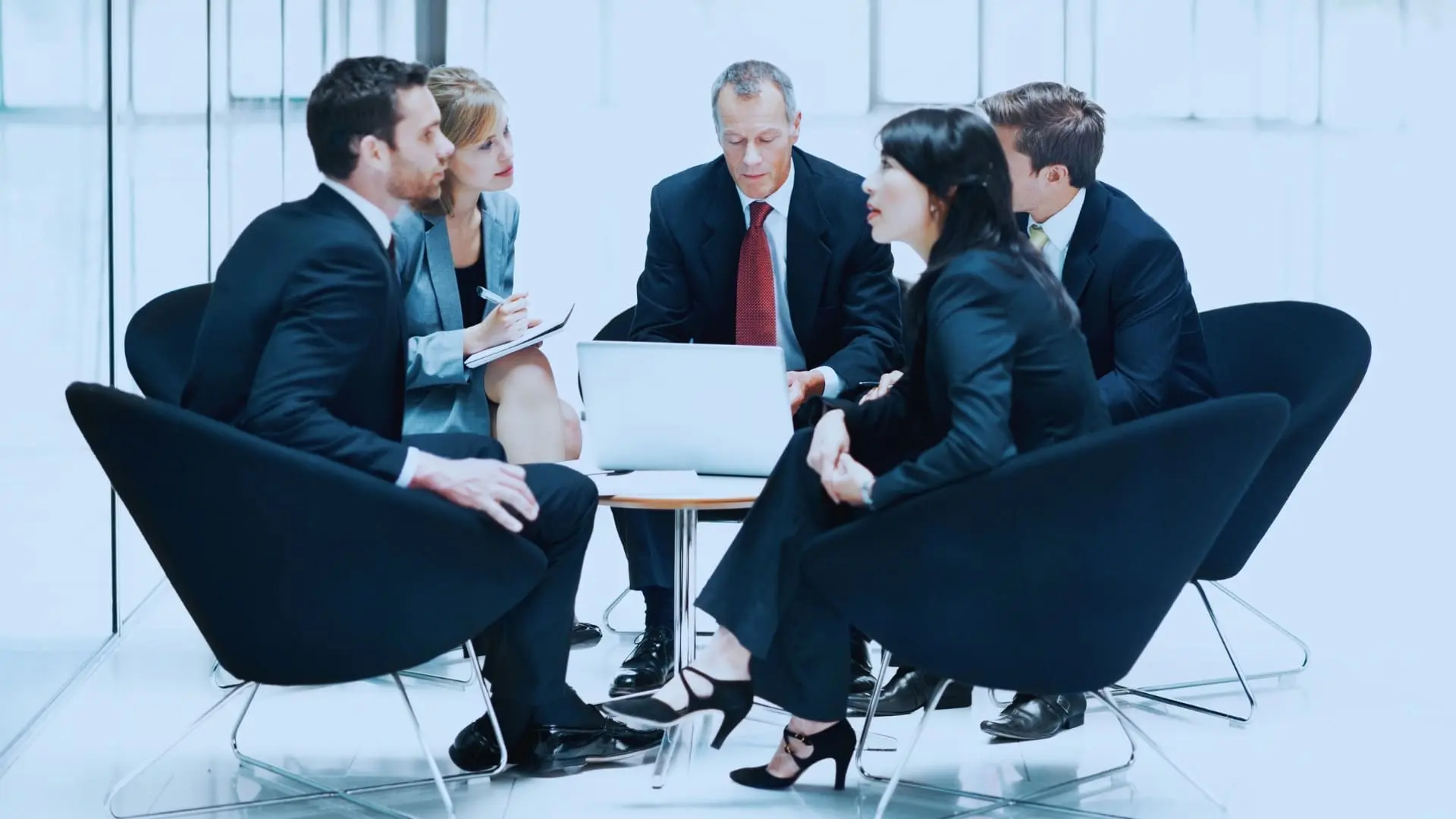Five business professionals in formal attire sit in a circle around a small table, engaged in a discussion comparing TeamGantt vs Monday.com, with a laptop open in the center in a modern, bright office space.