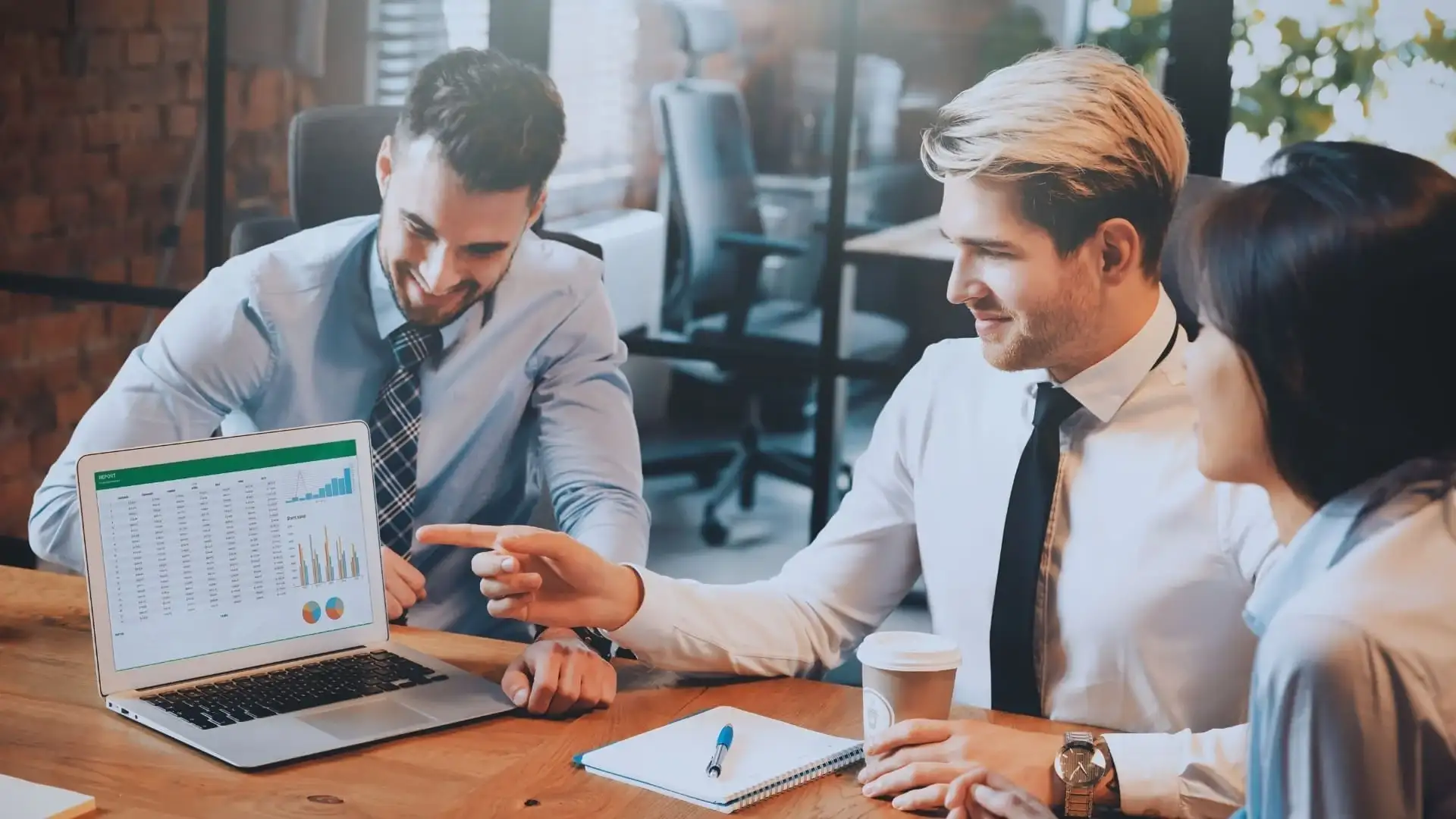 Three people in business attire sit at a table, looking at a laptop displaying TeamGantt vs Monday.com charts and graphs. One person points at the screen while another holds a coffee cup, engaged in a focused discussion or meeting.