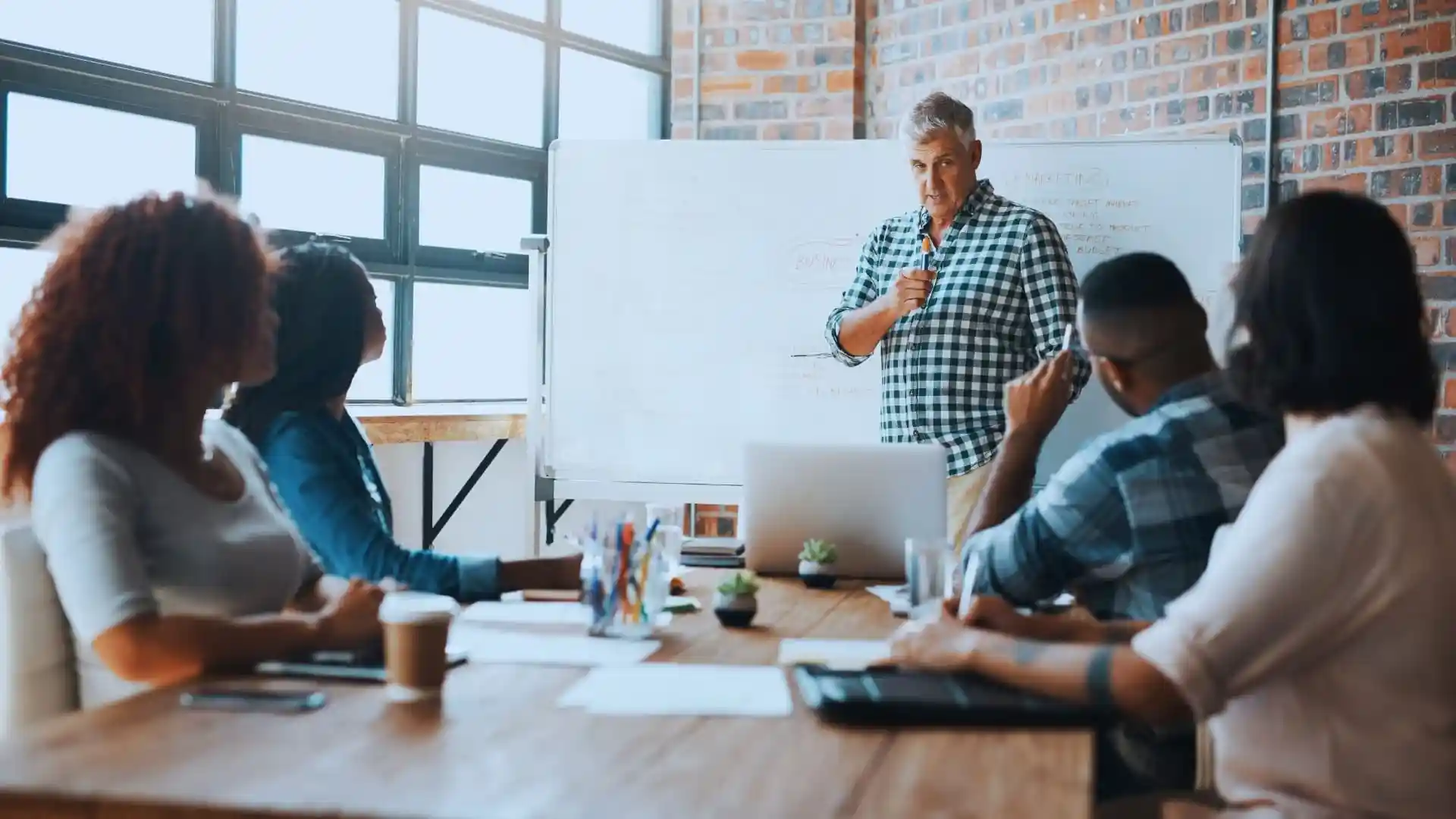 A man stands by a whiteboard giving a presentation on Monday.com Price to three seated colleagues in a modern office with large windows and exposed brick walls. Laptops, coffee cups, and notebooks are on the table.