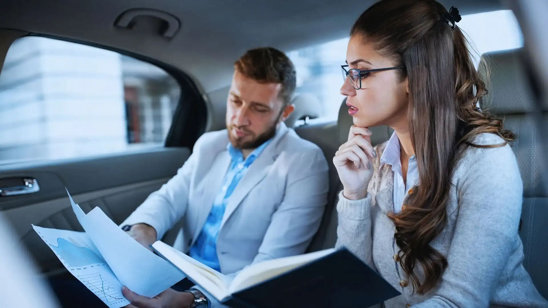 Two business professionals sit in the backseat of a car, reviewing documents together. As they compare notes on TeamGantt vs Monday.com, the man holds papers while the woman, wearing glasses, looks on thoughtfully.
