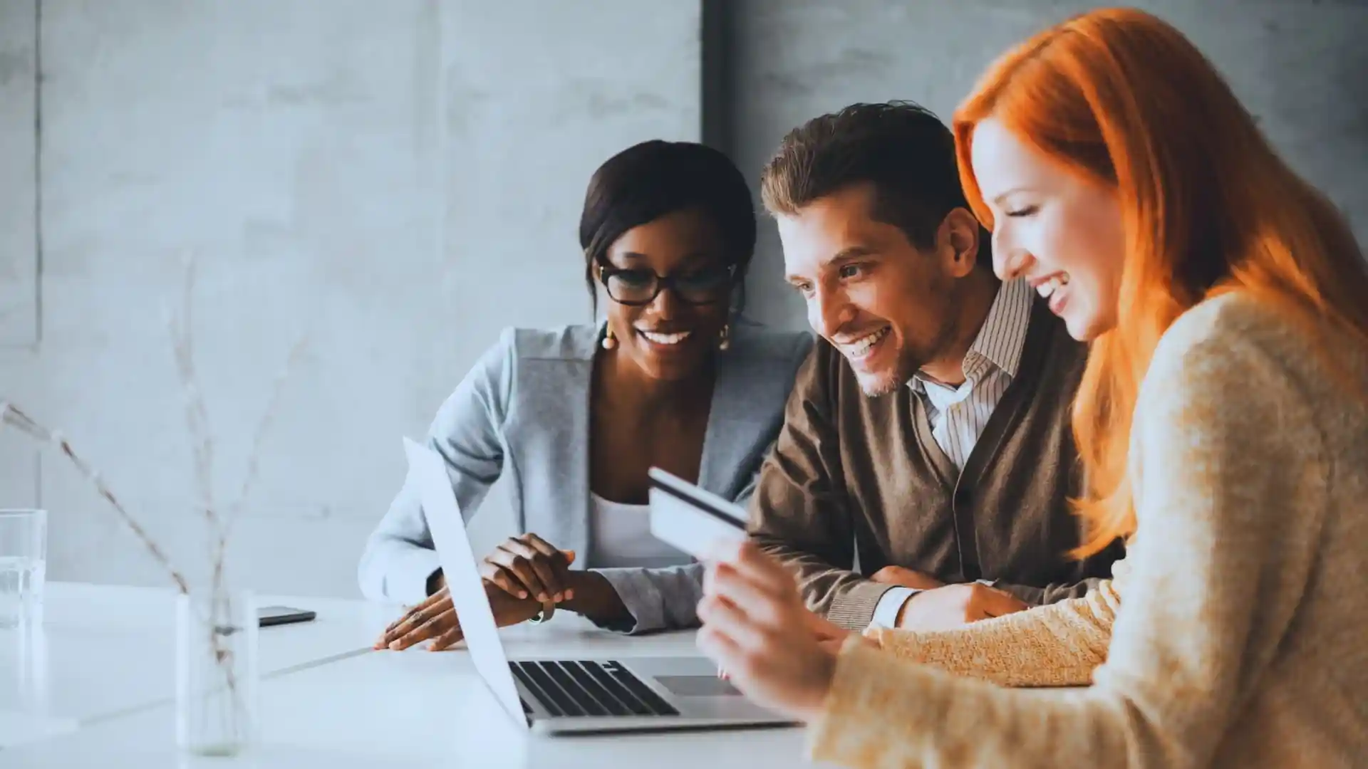 Three people sit at a table, smiling and looking at a laptop. One person holds a credit card, suggesting they are discussing an online purchase or reviewing Monday.com price options together.