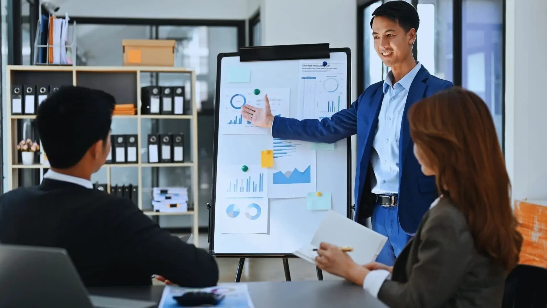 A man in a blue suit presents charts and graphs comparing TeamGantt vs Monday.com to two colleagues seated at a conference table in a modern office setting.