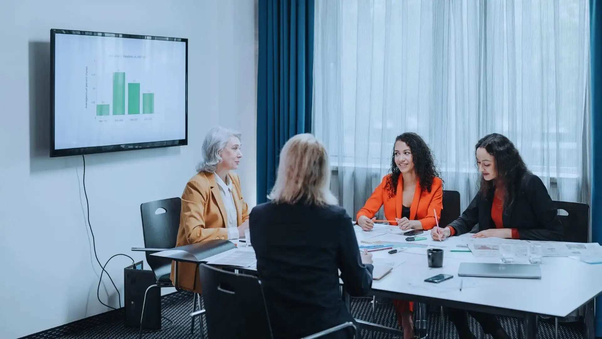 Four women in business attire sit around a conference table having a meeting with documents and laptops, discussing project management tools such as TeamGantt vs Monday.com. A presentation with a green bar graph is displayed on a screen on the wall.