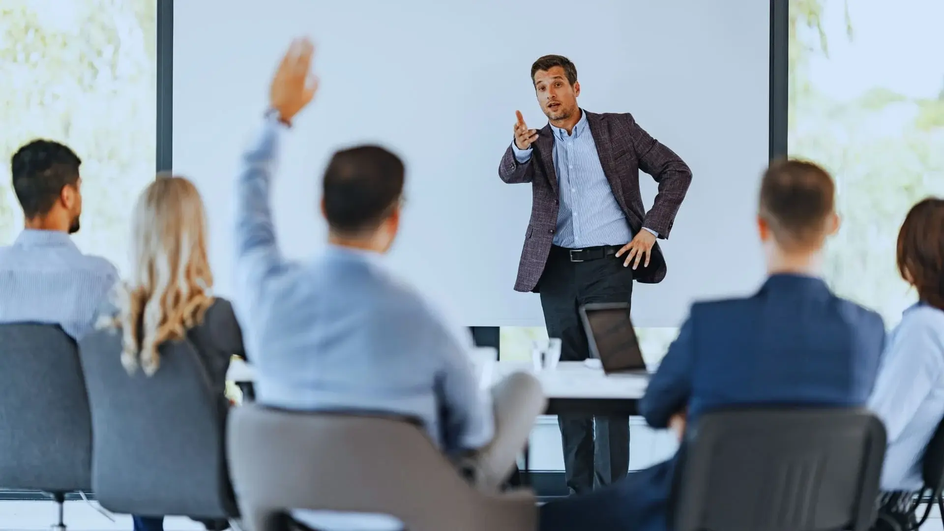 A man in a suit stands and speaks in front of a group during a meeting on TeamGantt vs Monday.com; one audience member raises their hand to ask a question.