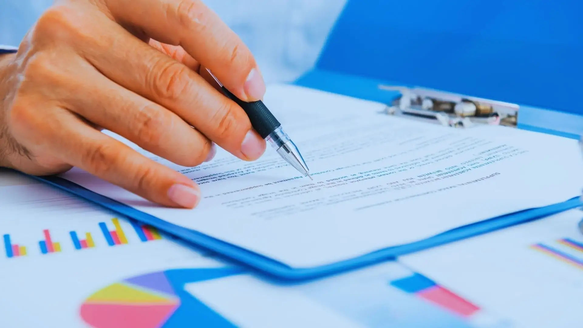 A person holding a pen reviews a document on a clipboard, with charts and graphs—perhaps comparing TeamGantt vs Monday.com—partially visible on the table in the background.