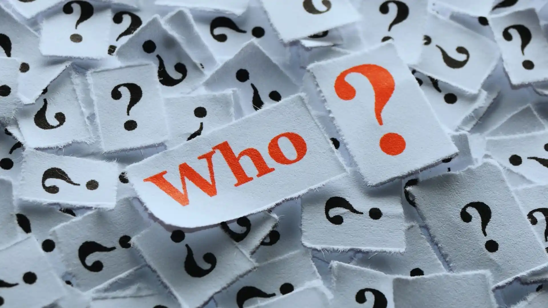 A pile of small paper pieces with black question marks. In the center, one piece stands out with the word "Who" in bold red letters, hinting at a decision—TeamGantt vs Monday.com?.