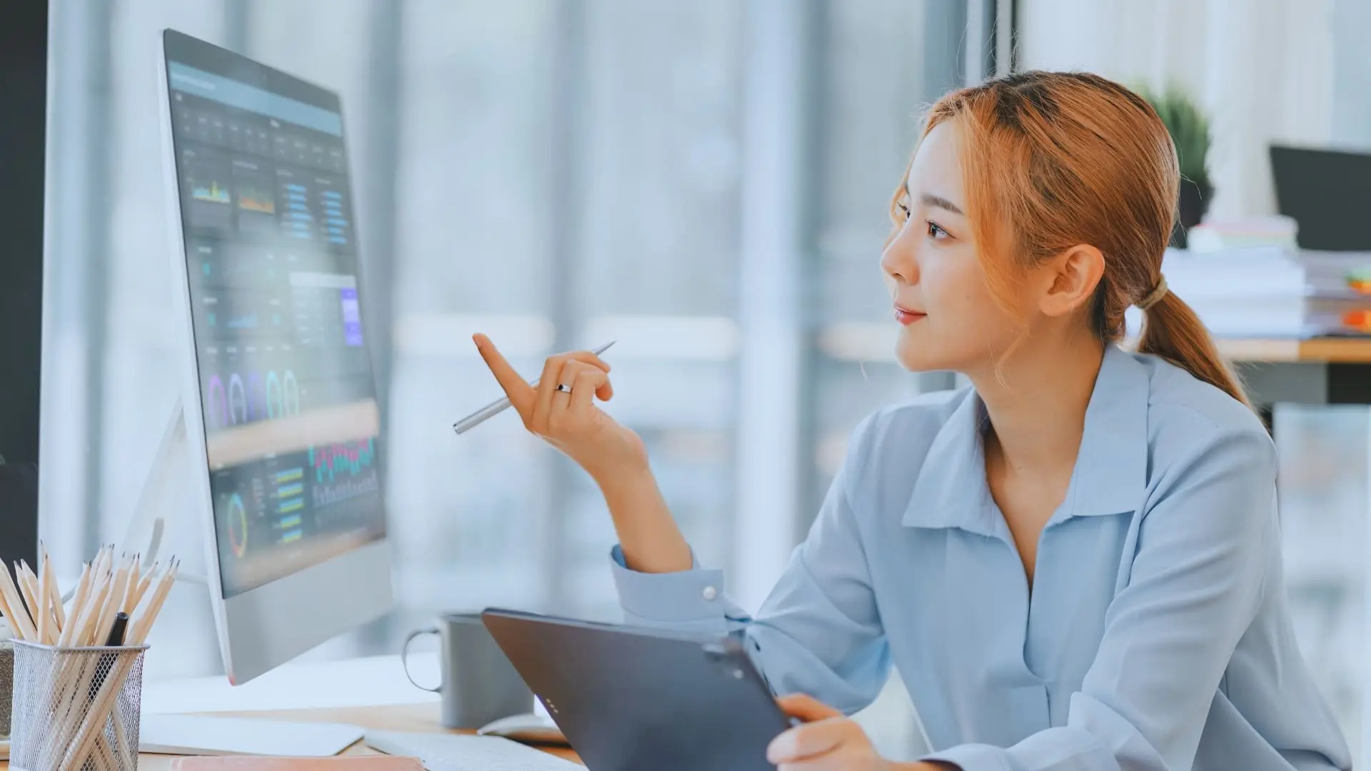 A woman in a light blue shirt sits at a desk, holding a digital tablet and stylus, reviewing Monday.com price charts displayed on a large computer monitor in a bright modern office.
