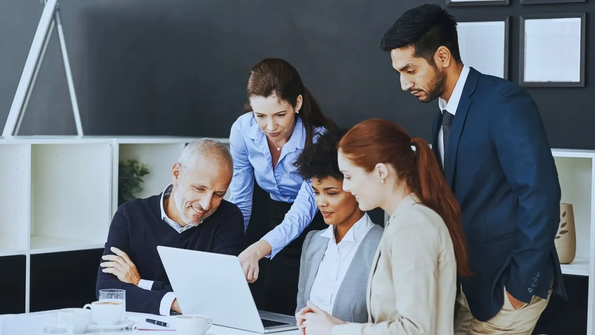 Five professionally dressed colleagues gather around a laptop in an office, collaborating and discussing Monday.com price options on the screen with interest and engagement.