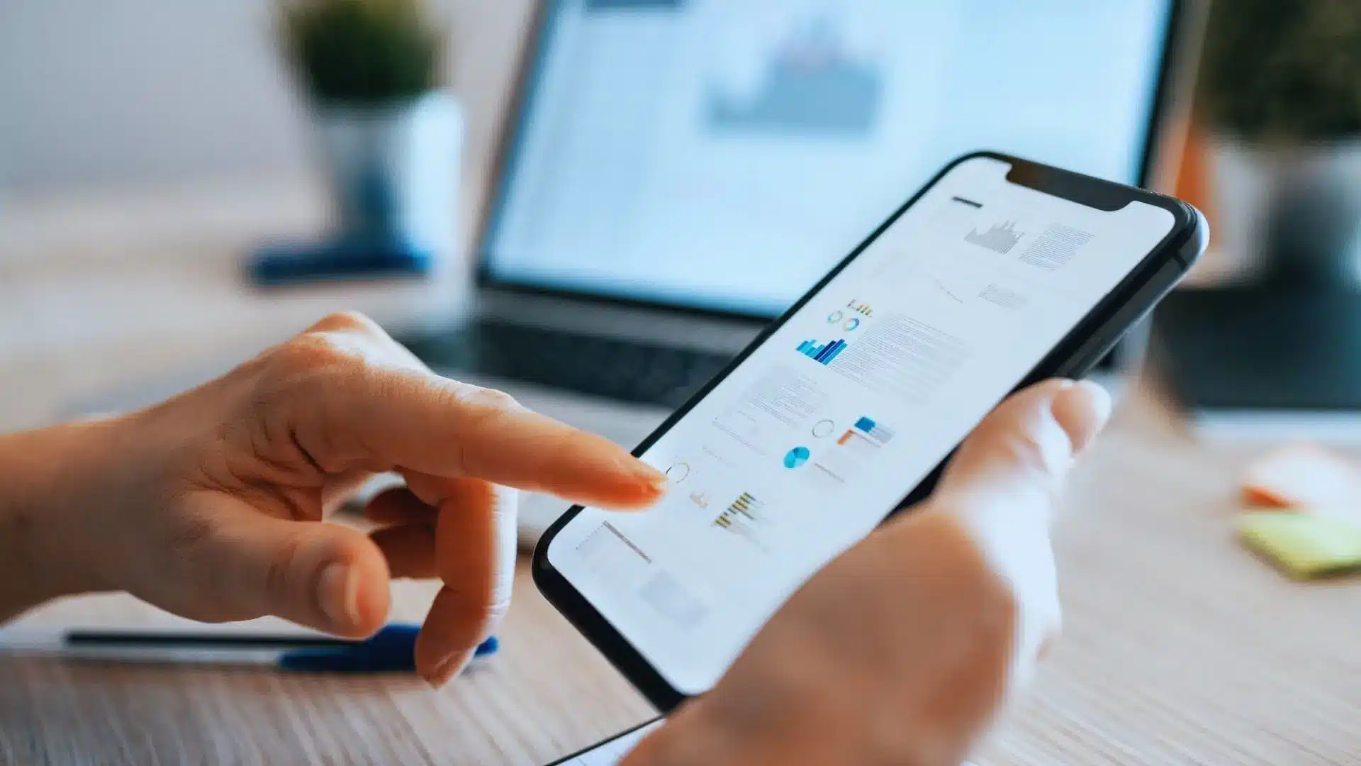 A person uses a smartphone to view graphs and documents, with a blurred laptop and office supplies in the background, suggesting multitasking or remote work—perhaps checking Monday.com price details on the go.