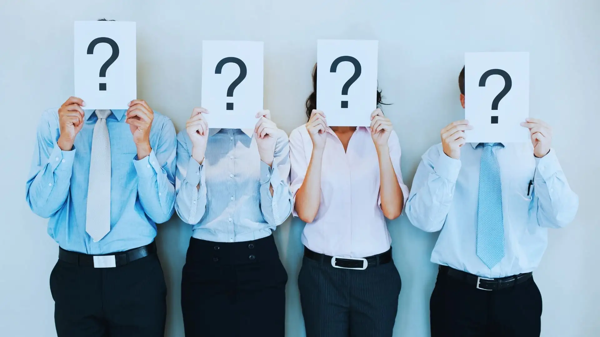 Four people in business attire stand side by side, each holding a sheet with a large question mark that hides their face. Against a plain background, they represent the unknowns you might face—like finding the right Monday.com price for your team.